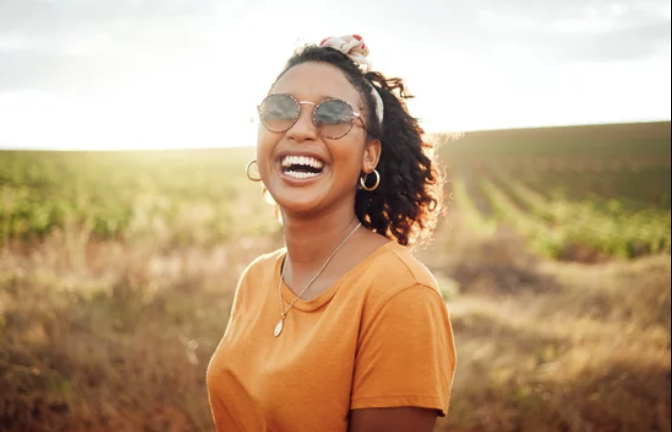 A woman with curly hair wearing sunglasses, hoop earrings, necklace, and an orange T-shirt, smiling outdoors in a field during sunset.
