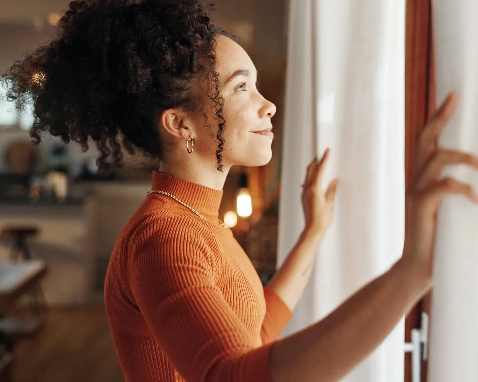 A woman with curly hair wearing a burnt orange turtleneck sweater, gold hoop earrings, and a gold chain necklace, standing by a window with white curtains and gently touching the curtain with her hands, looking outside with a slight smile.