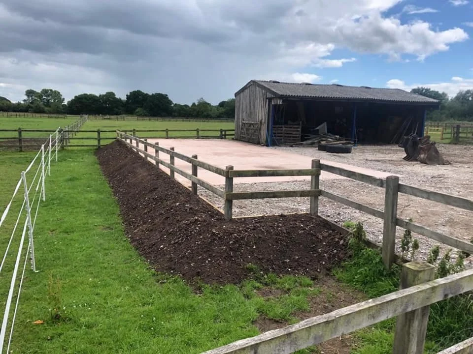 A farmyard with a newly constructed concrete pad, wooden fencing, and an old barn in the background under a partly cloudy sky.