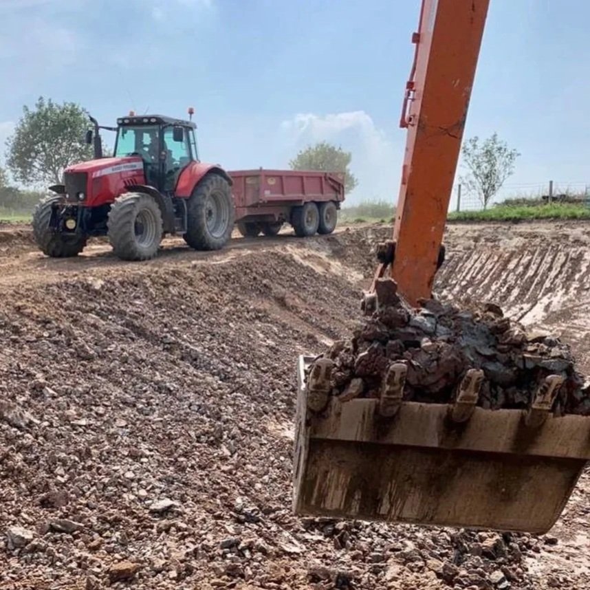 A construction site with a red tractor pulling a trailer, and an excavator bucket filled with dirt and rocks.