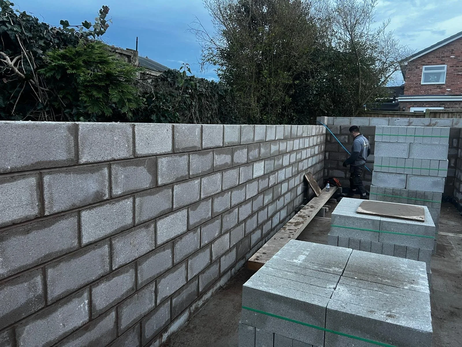 A worker installing concrete blocks to build a wall outdoors, with additional stacks of blocks nearby, under a partly cloudy sky.