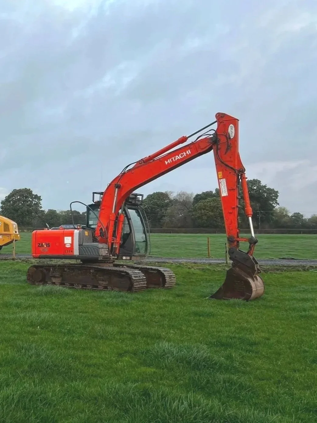 Orange Hitachi excavator on a grassy field with trees in the background and a cloudy sky.