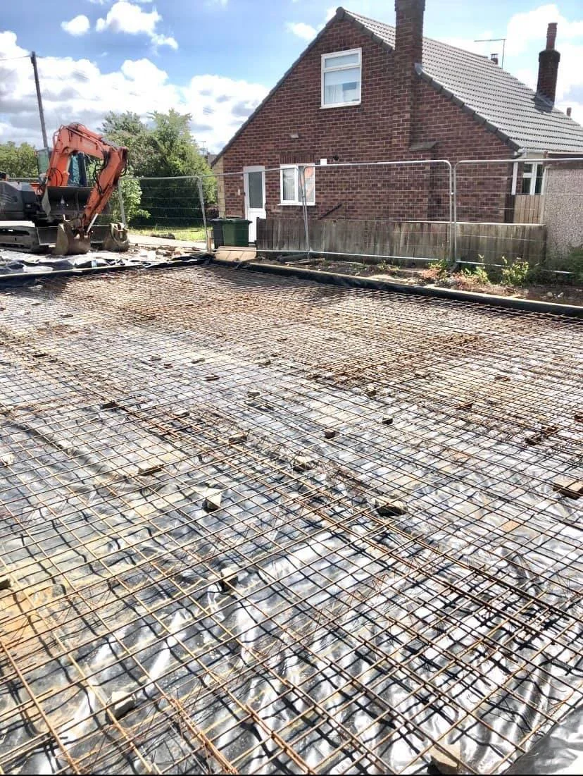 Construction site with rebar grid and excavator, house in background, partly cloudy sky.