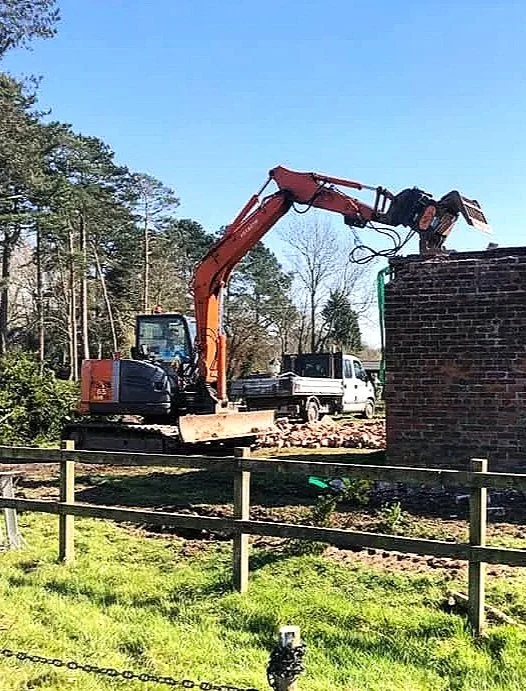 A small orange excavator is breaking a brick wall with its attachment, located outdoors near trees, with a grassy area and a wooden fence in the foreground.