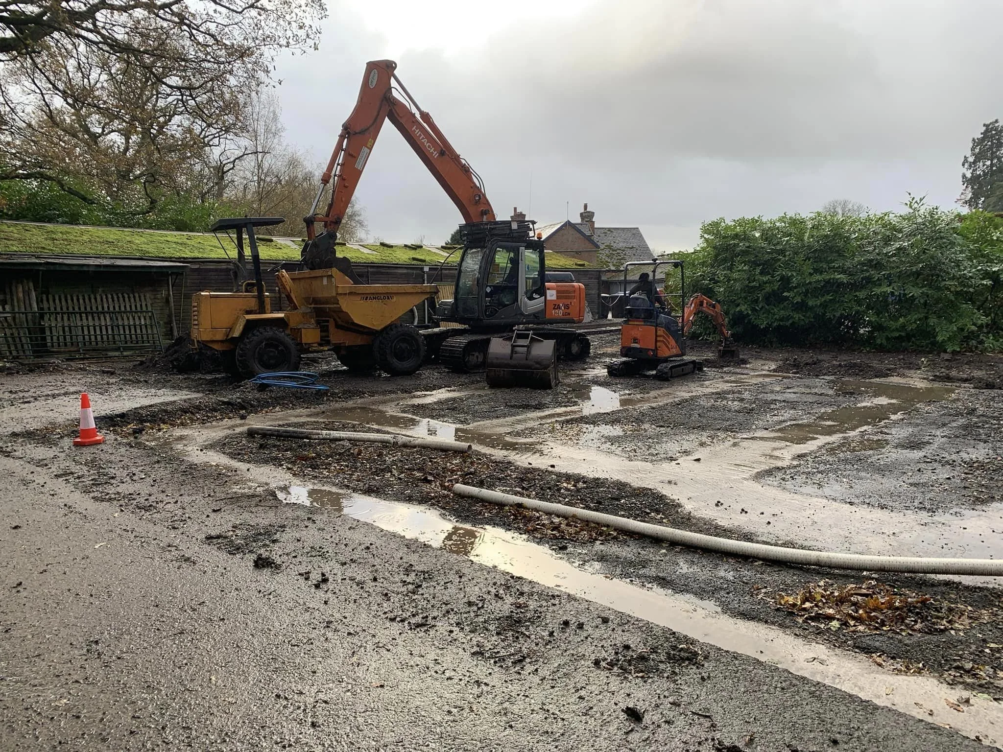 Construction site with machinery including an excavator and a dump truck, muddy ground, and a background of trees and houses with overcast sky.
