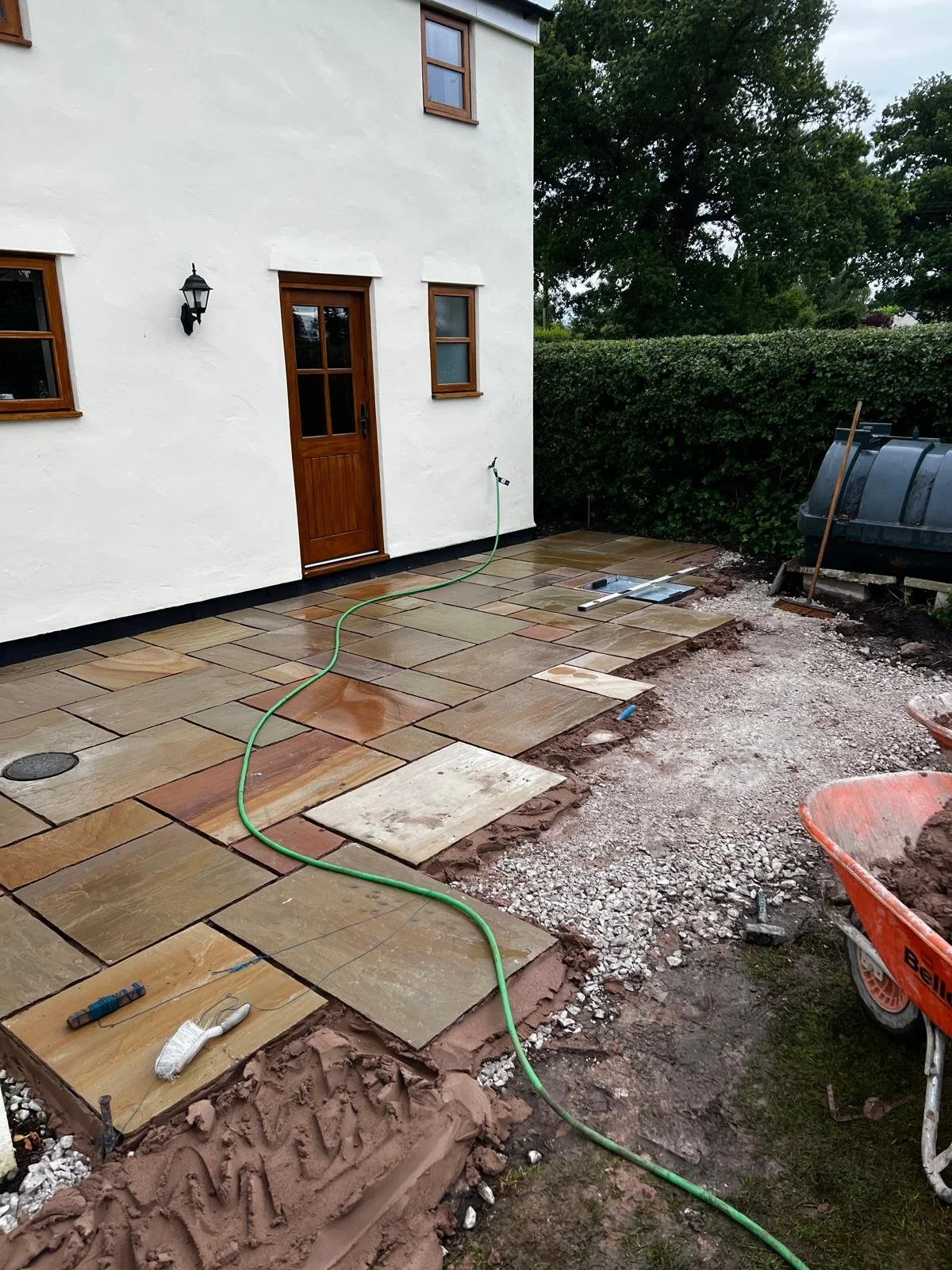 Backyard of a house with a newly installed stone patio, construction tools, and a water hose. The house has white walls, wooden-framed windows, and a door with a black outdoor lantern. There are green bushes and trees in the background.