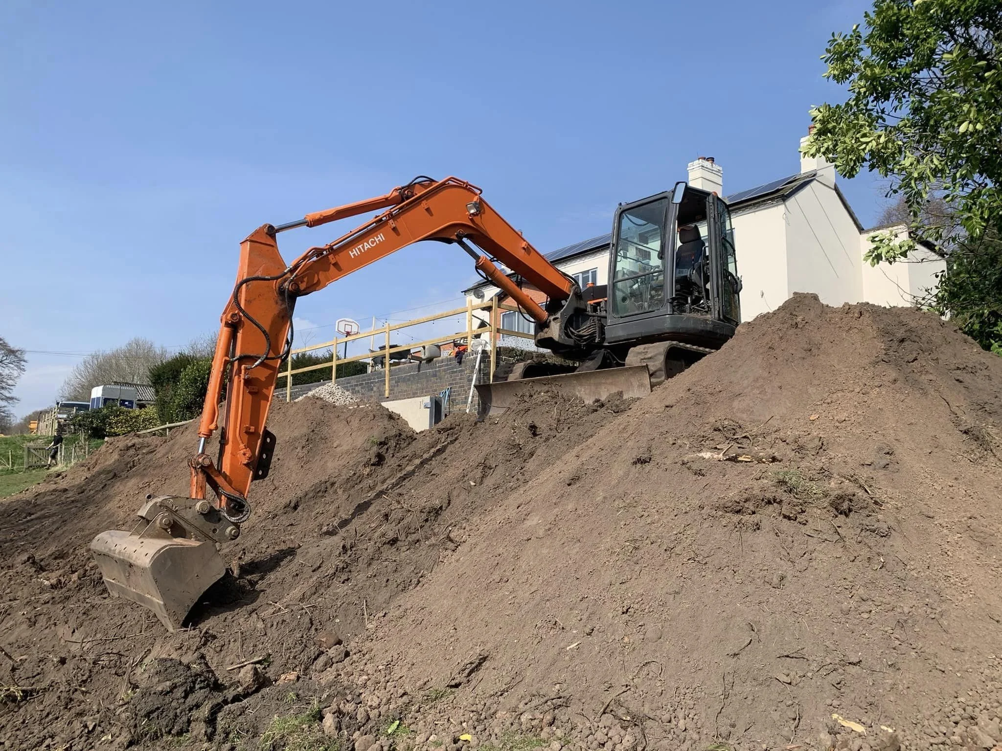 An orange Hitachi excavator digging into a large pile of dirt outdoors on a clear day, with a house, trees, and a basketball hoop visible in the background.