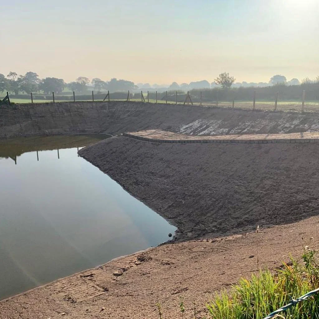 Construction site with a large, excavated pond or reservoir, surrounded by a dirt embankment and a wooden fence beyond, at sunrise or sunset with a hazy, peaceful landscape in the background.