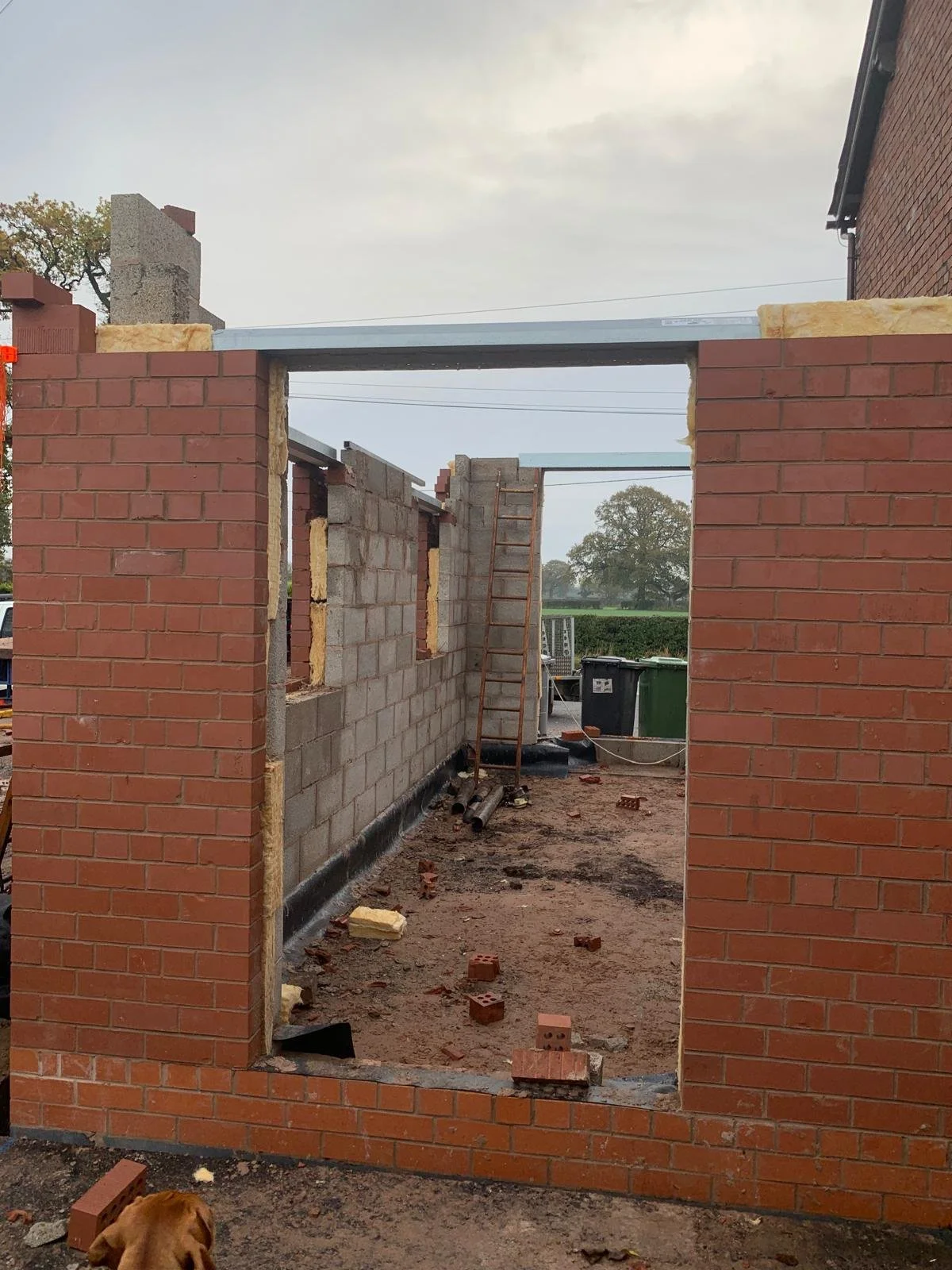Construction site with brick walls being built, a ladder leaning against the wall, construction materials and trash on the ground, overcast sky.