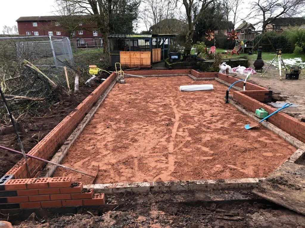 A backyard under construction with a new brick foundation, construction tools, and garden decorations in the background.
