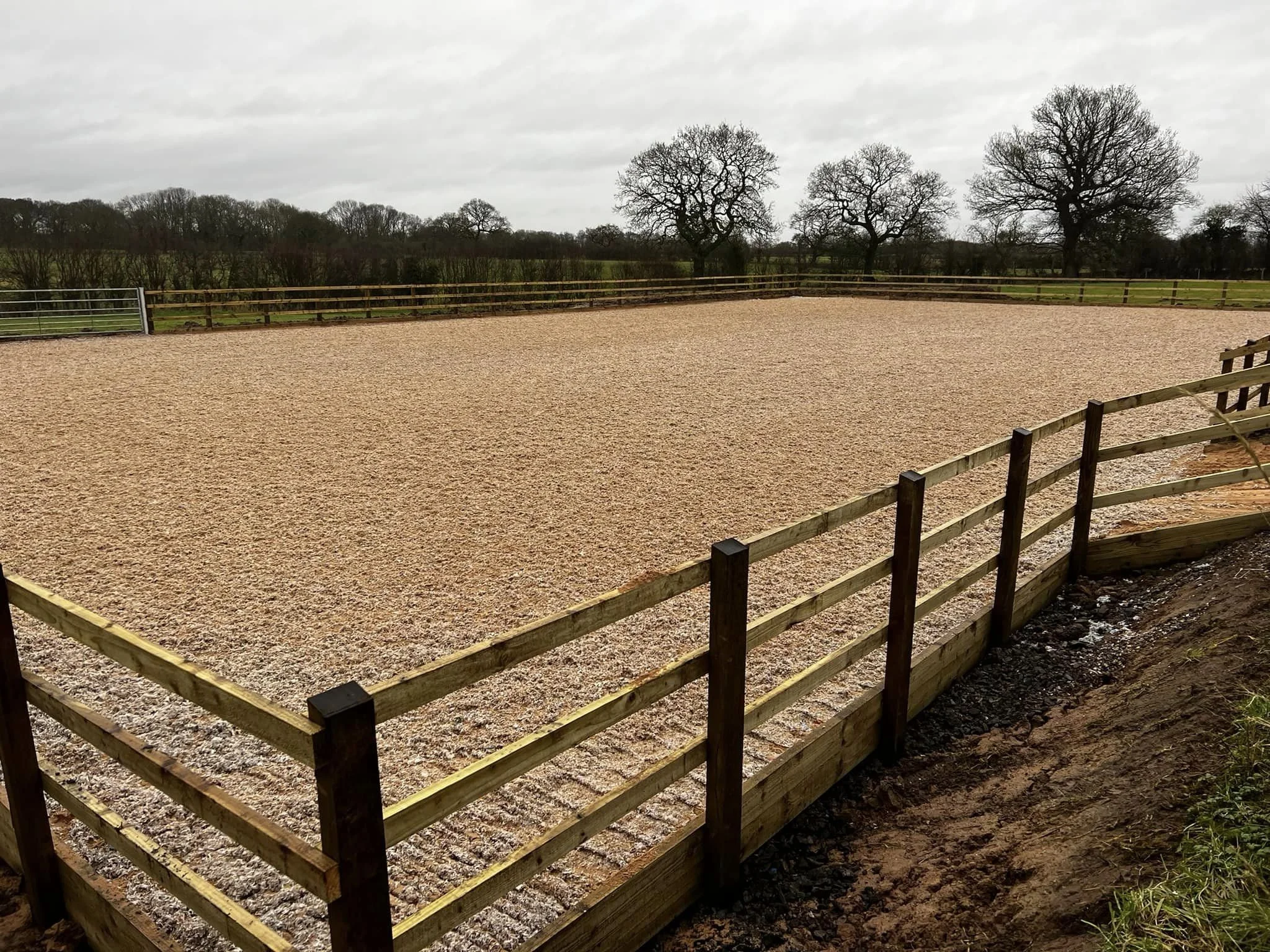 An outdoor arena with a gravel surface surrounded by a wooden fence, with leafless trees and a cloudy sky in the background.