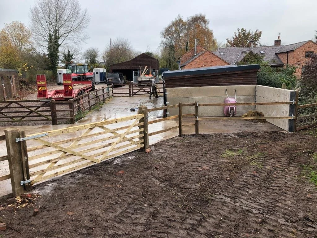 A muddy outdoor farm or rodeo pen with wooden and concrete fencing, a pink wheelbarrow, and various farm equipment, with houses and trees in the background on a cloudy day.