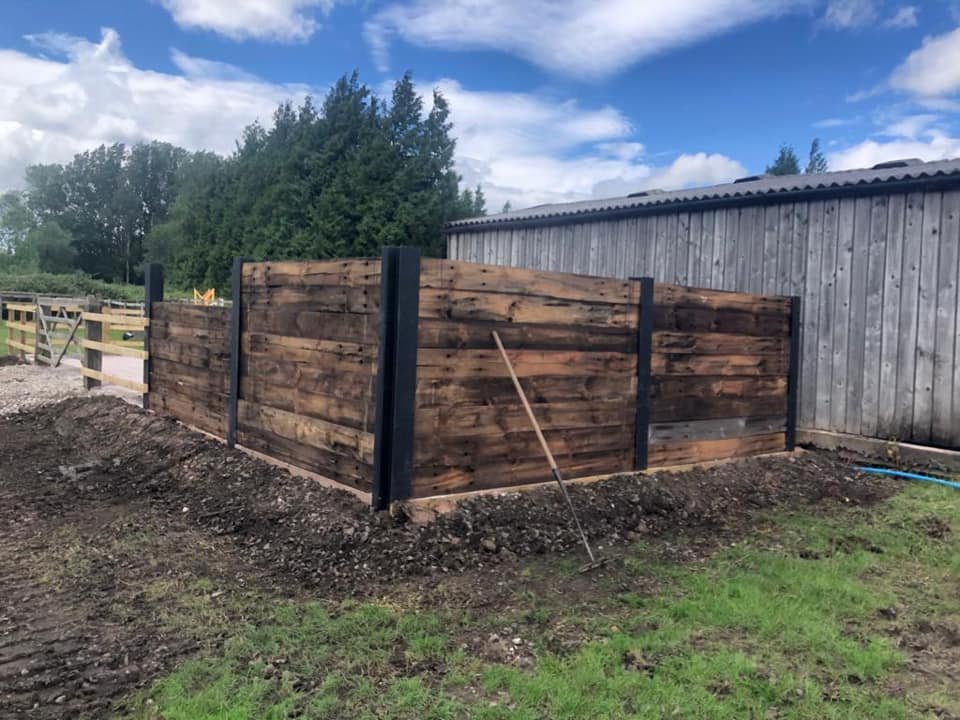 A wooden privacy fence with black posts next to a weathered gray shed, set in a grassy and dirt area under a partly cloudy sky.