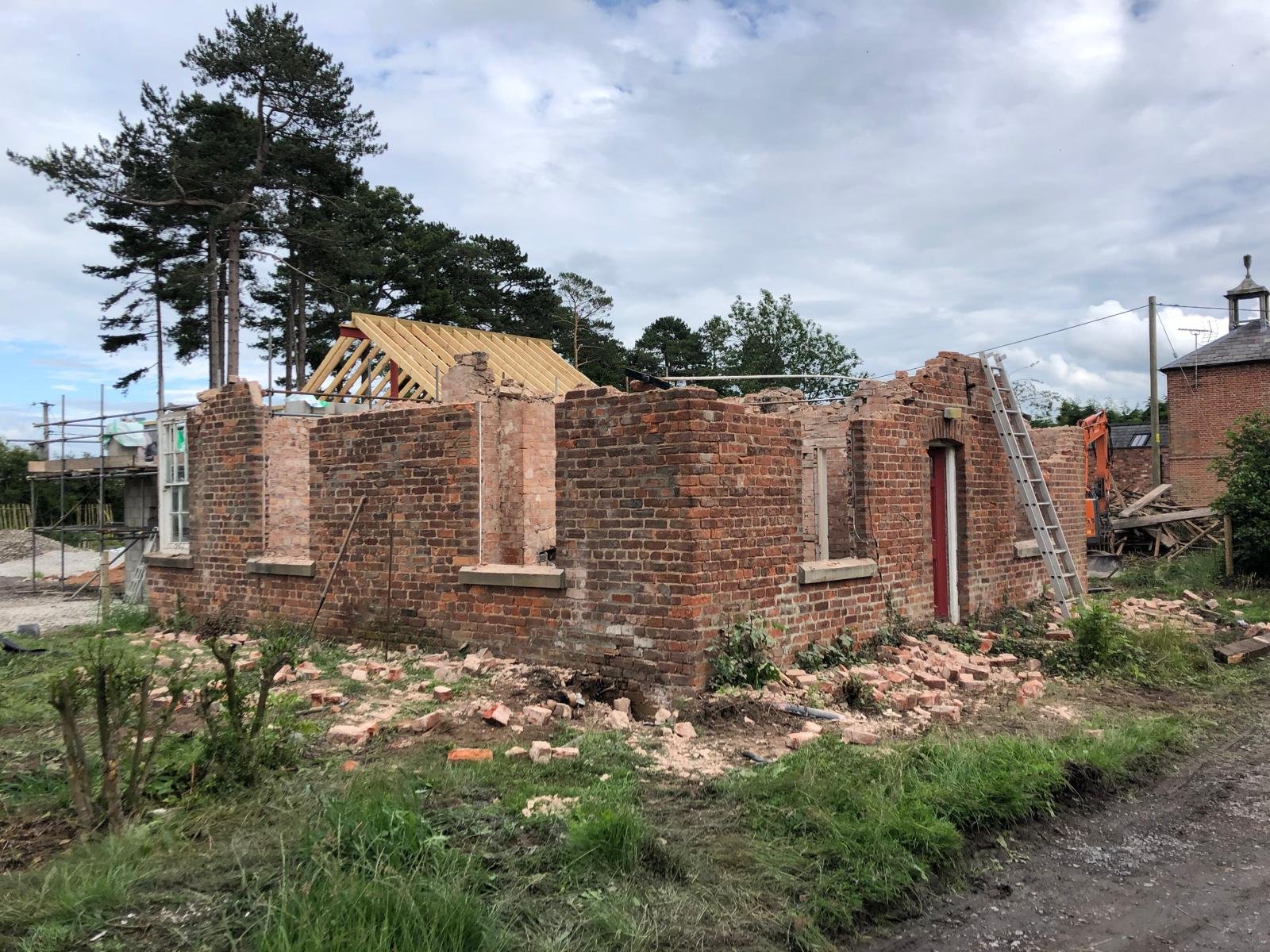 A brick building under renovation with partially removed roof framing, a ladder leaning against the wall, construction equipment, and scattered bricks on the ground.