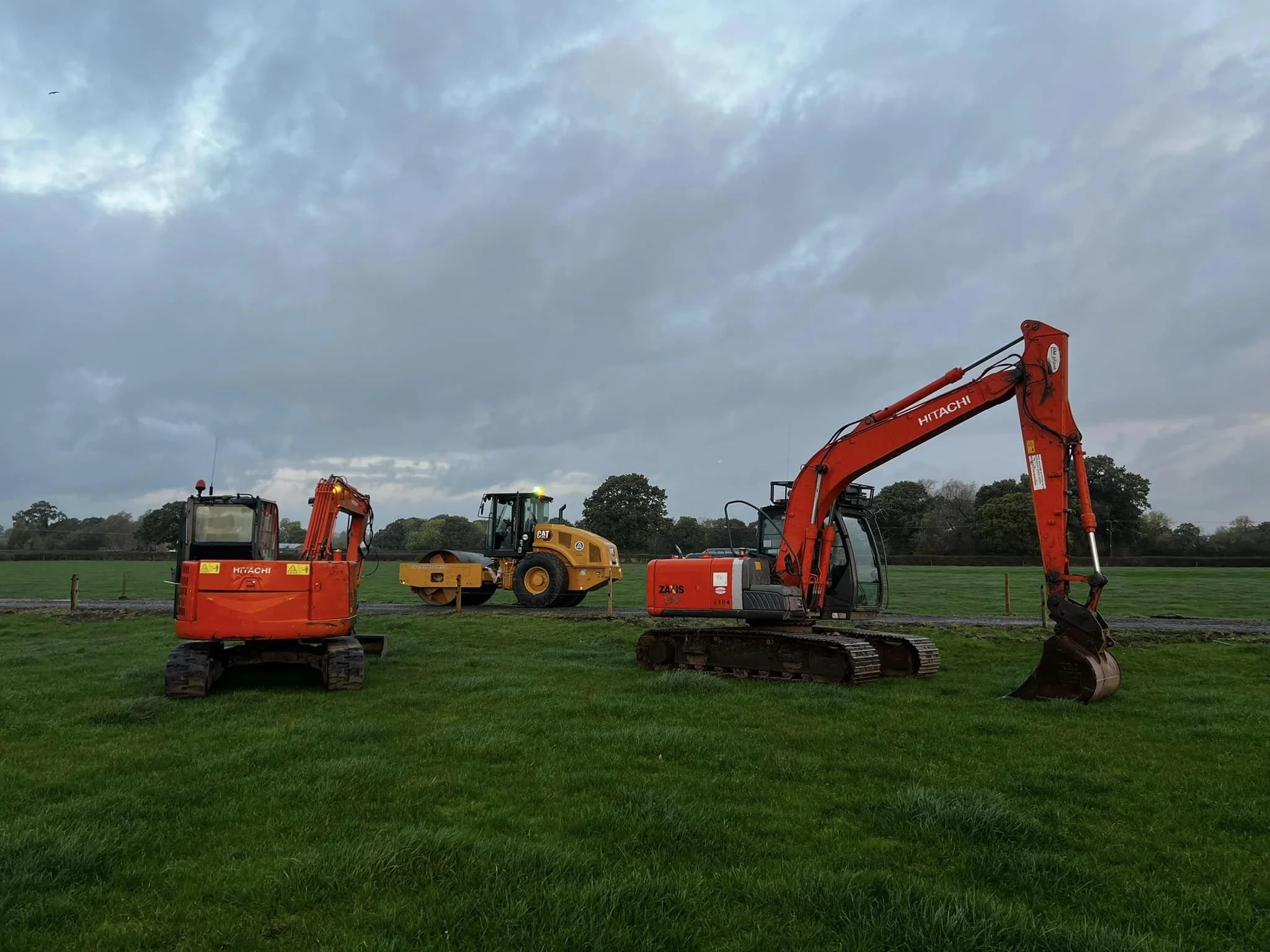 Construction equipment, including a bright orange Hitachi excavator, yellow bulldozer, and other machinery, are positioned on a grassy field under a cloudy sky.