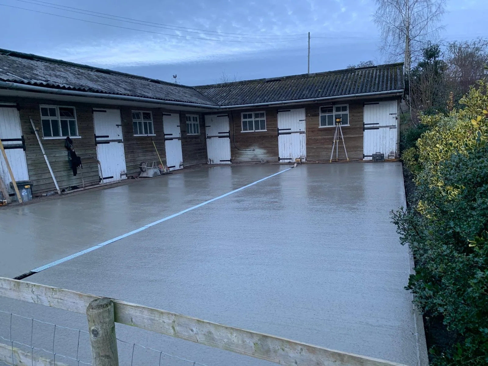 A wet concrete surface in front of a long wooden shed with multiple doors, some of which are painted white, on a cloudy day with trees and bushes on the right side.