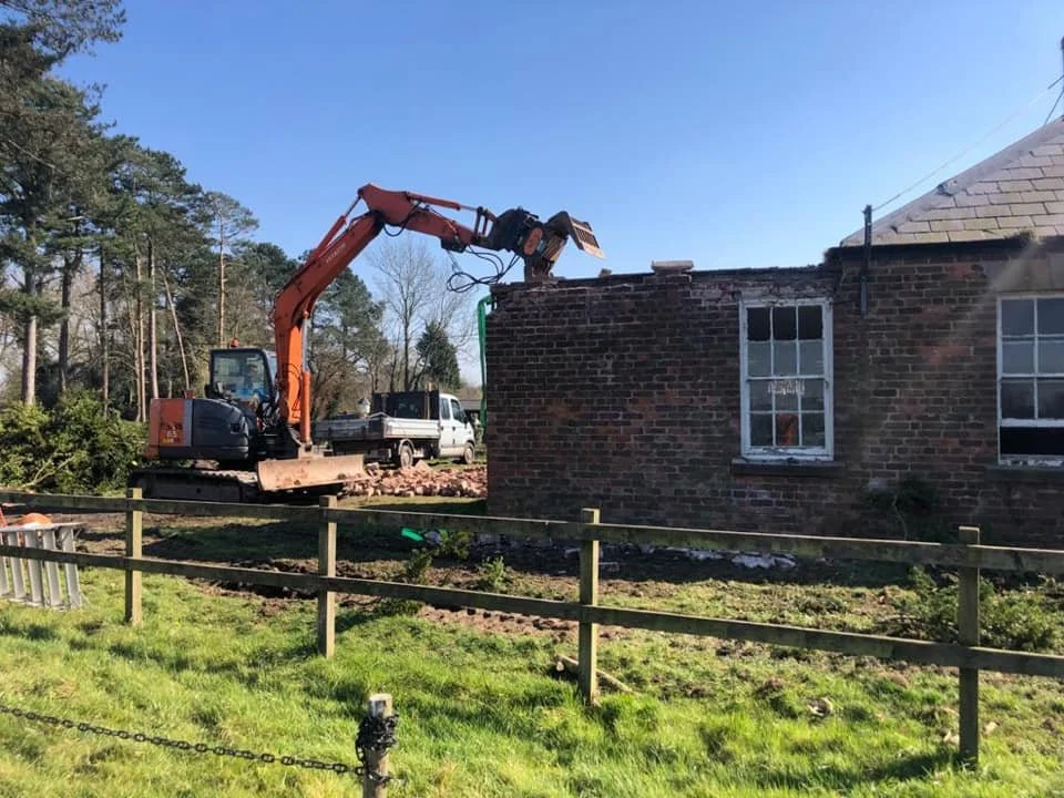 A construction scene with an excavator tearing down a brick building, with a wooden fence and grass in the foreground and a clear blue sky above.