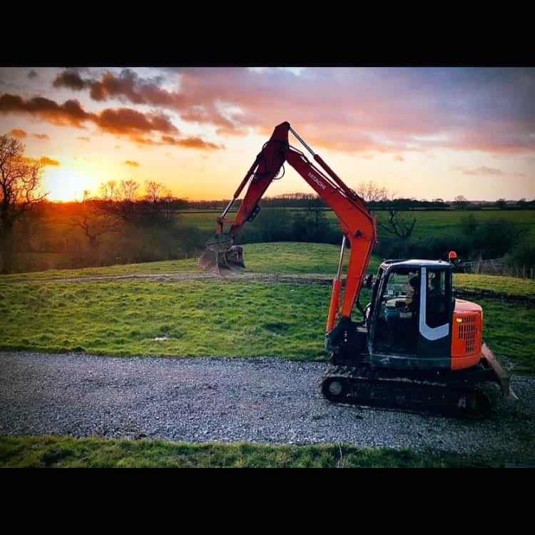 An orange and black excavator on a gravel path, digging into a grassy field at sunset with orange and purple clouds and trees in the background.