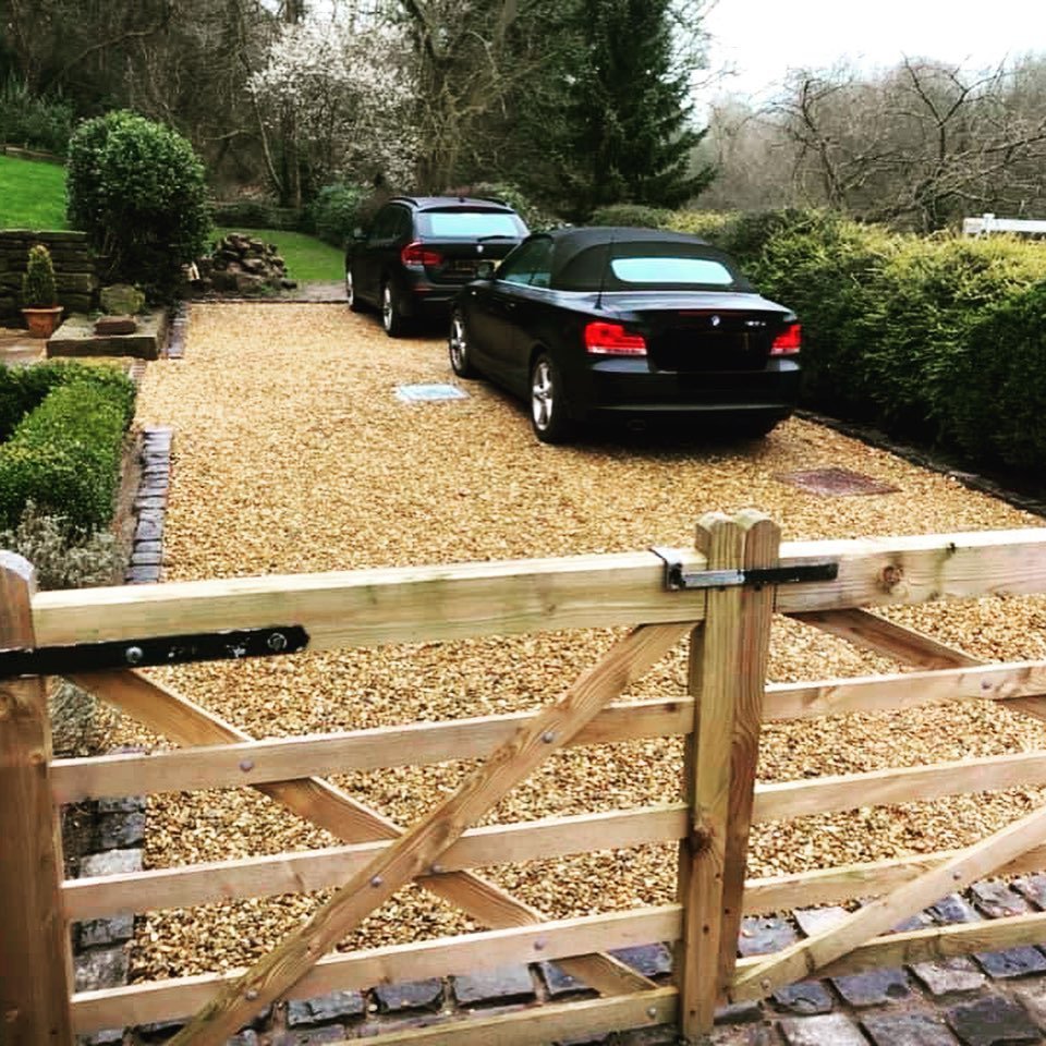 View of a driveway with two parked black cars, a black convertible in front, and an SUV behind, gated by a wooden fence, with bushes and trees in the background.
