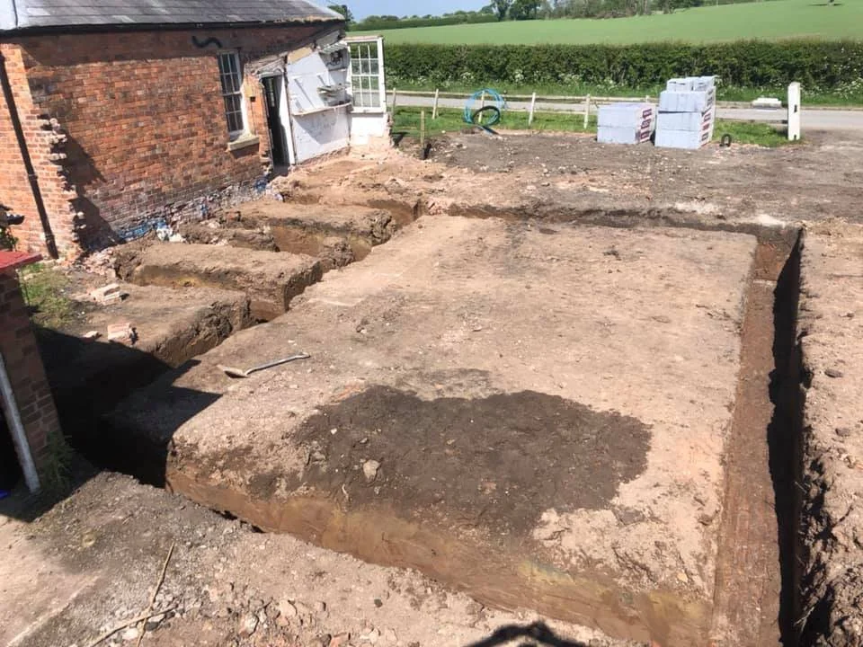 Excavated foundation trenches for a building on a construction site next to a brick house, with construction materials and tools nearby.