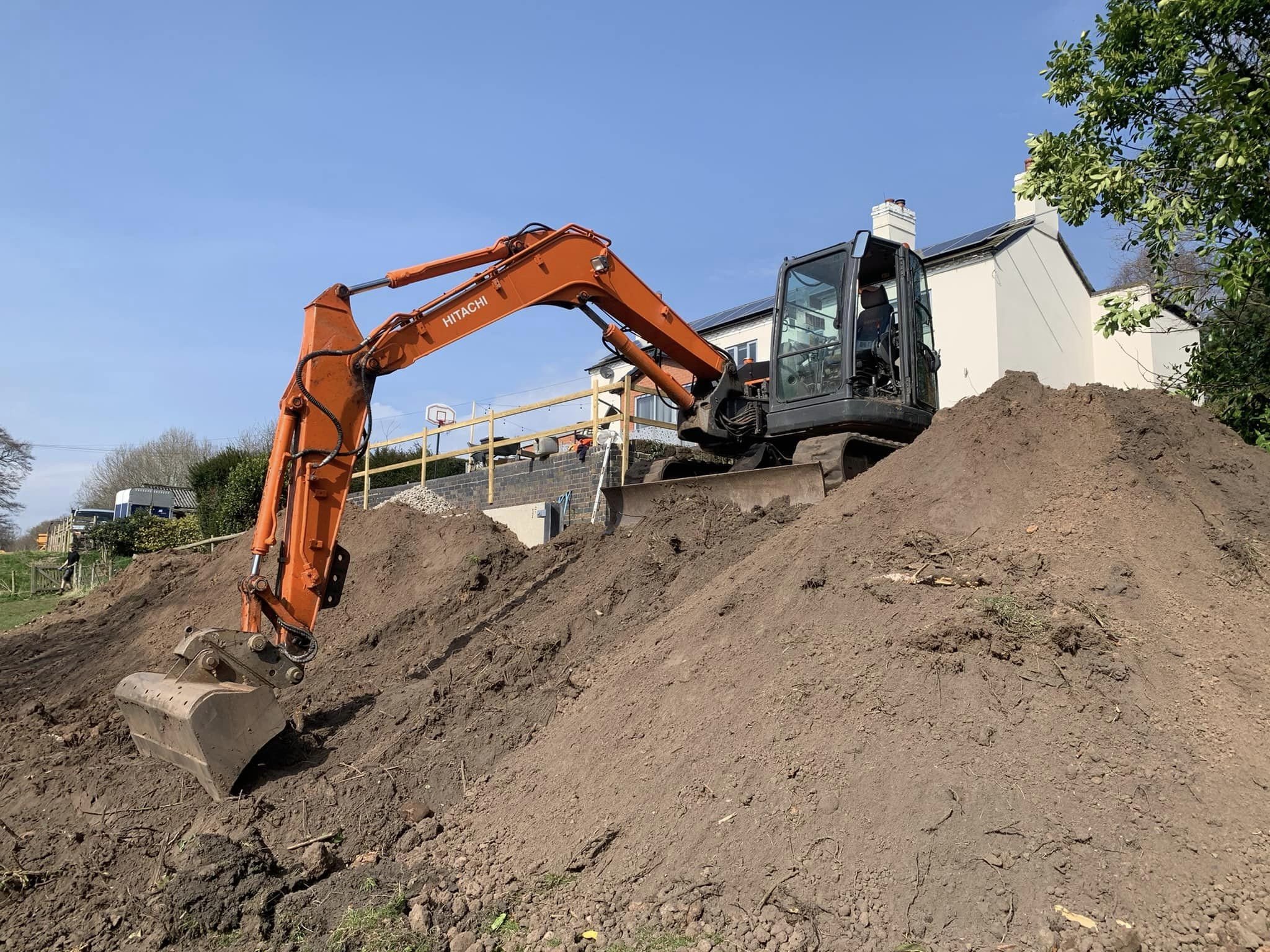 Construction site with an orange excavator digging into dirt, house with solar panels, and a clear blue sky.