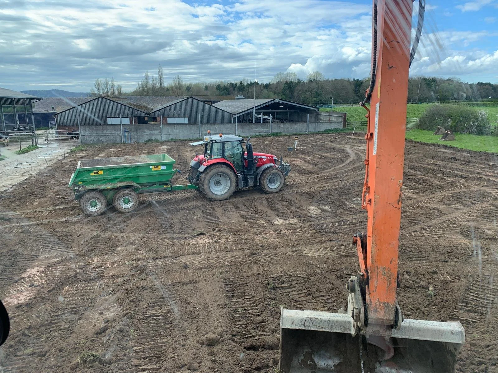 A construction site with a red tractor pulling a green trailer, surrounded by freshly tilled earth. An excavator arm is partially visible on the right side of the image. In the background, there are farm buildings and a partly cloudy sky.