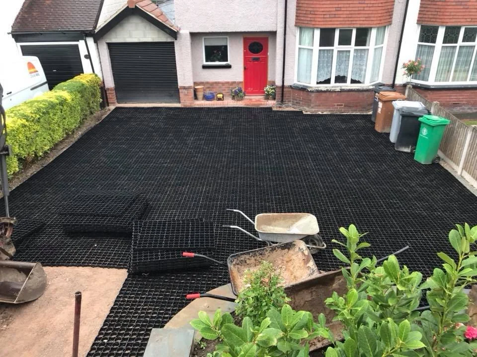 A backyard with a freshly installed black grid-based paving surface, with wheelbarrows, construction tools, and potted plants, in front of a house with a red door and large bay windows.