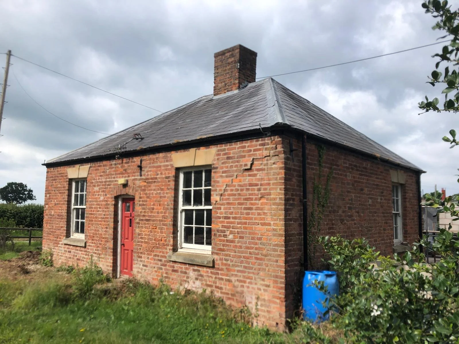 A small brick house with a red door and three windows, showing some cracks in the brick wall, under cloudy skies.