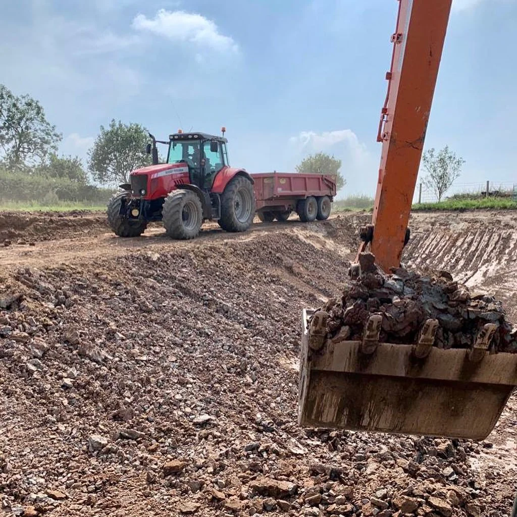 A red tractor with a trailer attached operates on a dirt construction site, with an excavator moving rocks and dirt in the foreground and a partly cloudy sky above.