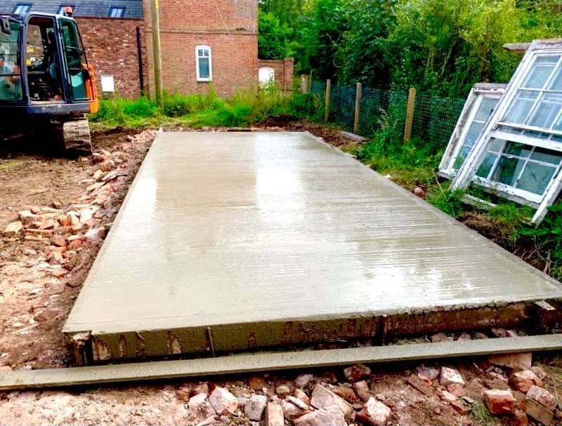 Freshly poured concrete slab on a construction site, with a excavator on the left and windows on the right.