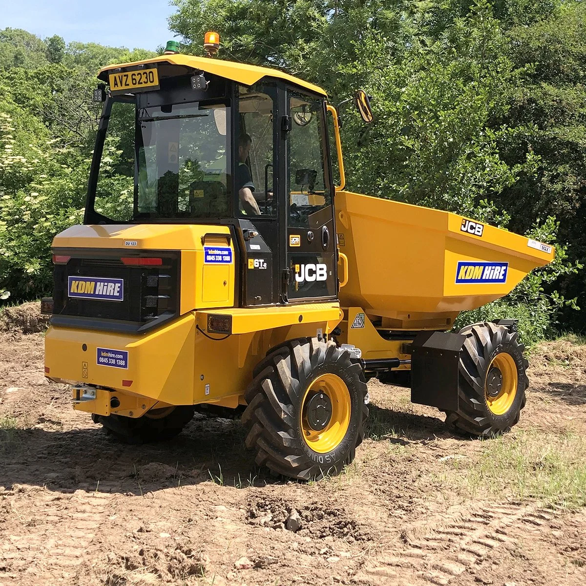 A yellow JCB dumper truck with KDM Hire branding on the side, parked on dirt with green trees in the background.