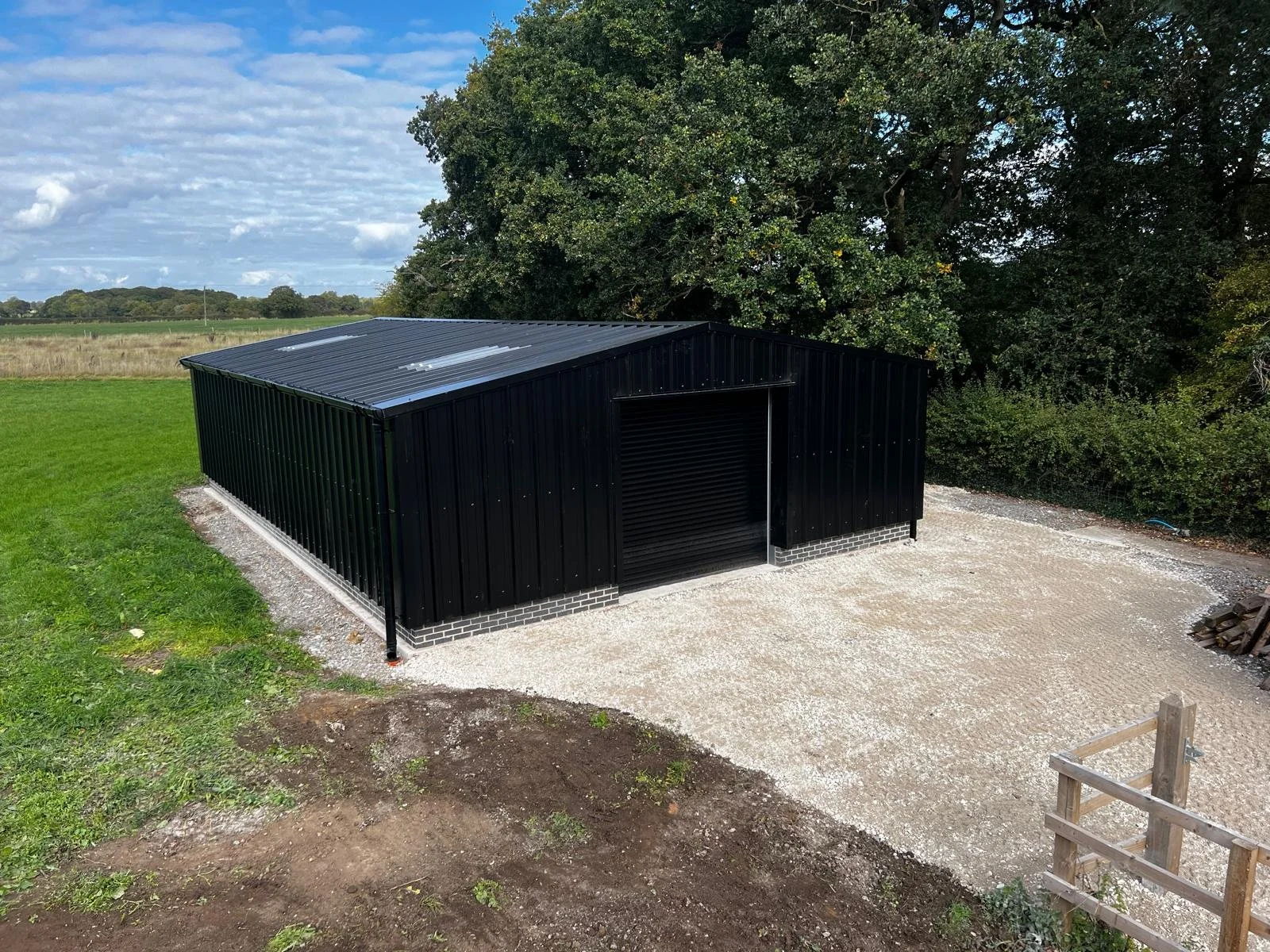 A black metal shed with a roller door, situated on a gravel area, with trees in the background and a partly cloudy sky overhead, in a rural setting.