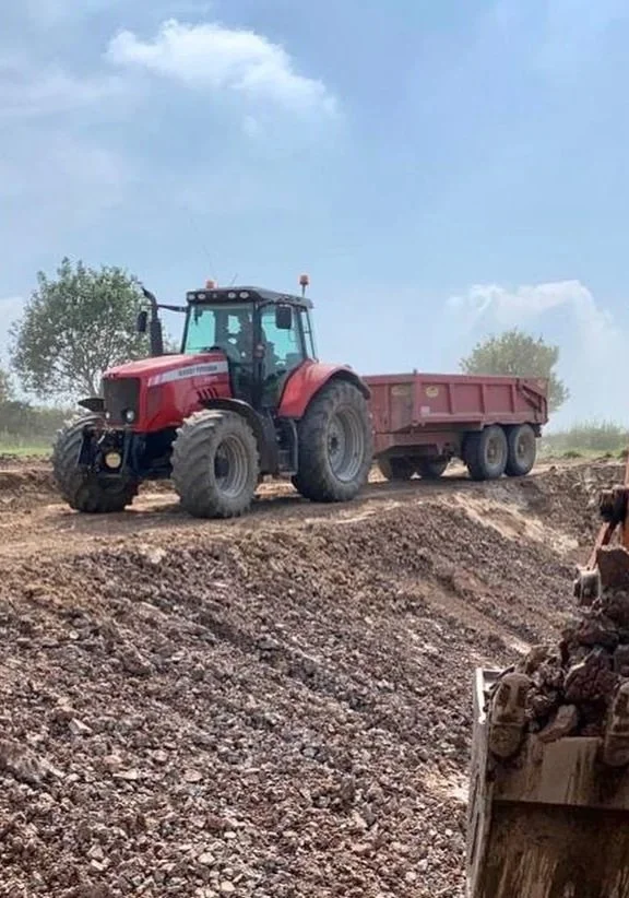 A red tractor with a trailer attached, parked on a dirt road or construction site with a cloudy sky and trees in the background.