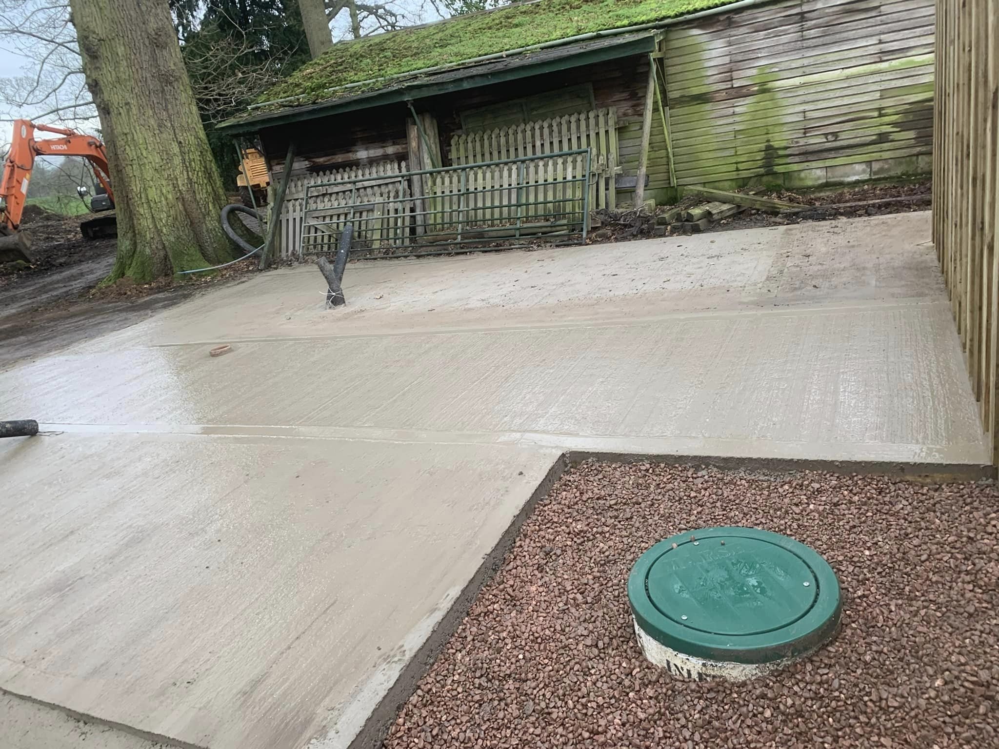 A construction site showing a concrete pathway, a fenced area with trees and an old wooden shed with moss on the roof. There's construction equipment partially visible on the left.