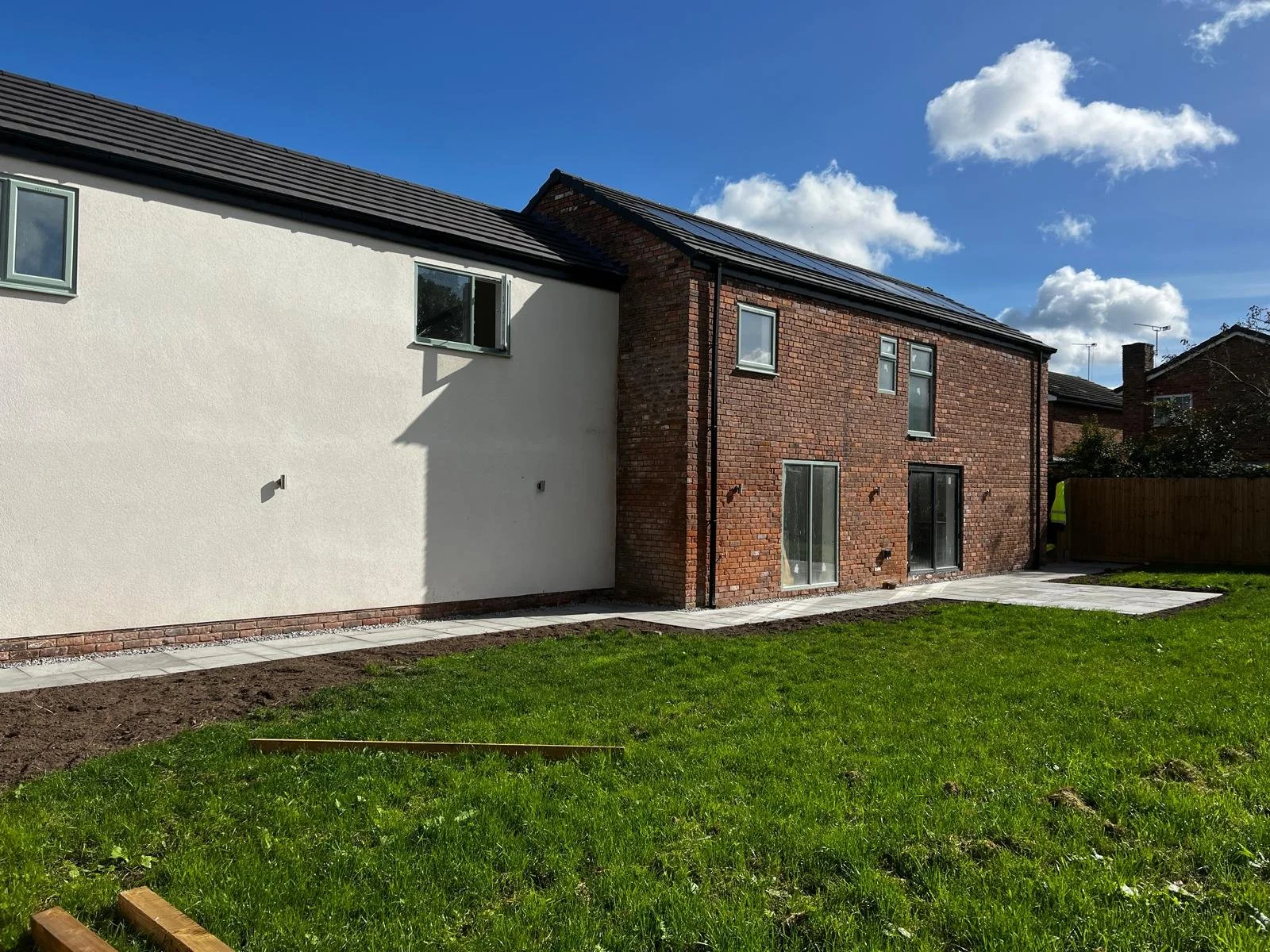 Newly constructed modern house with a white section and a red brick section featuring multiple windows and sliding glass doors, backyard with a green lawn, paved patio, and a wooden fence, under a partly cloudy sky.