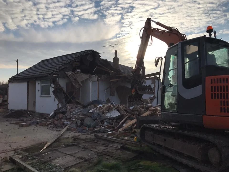 A house being demolished with an excavator during daylight, with debris and rubble on the ground.