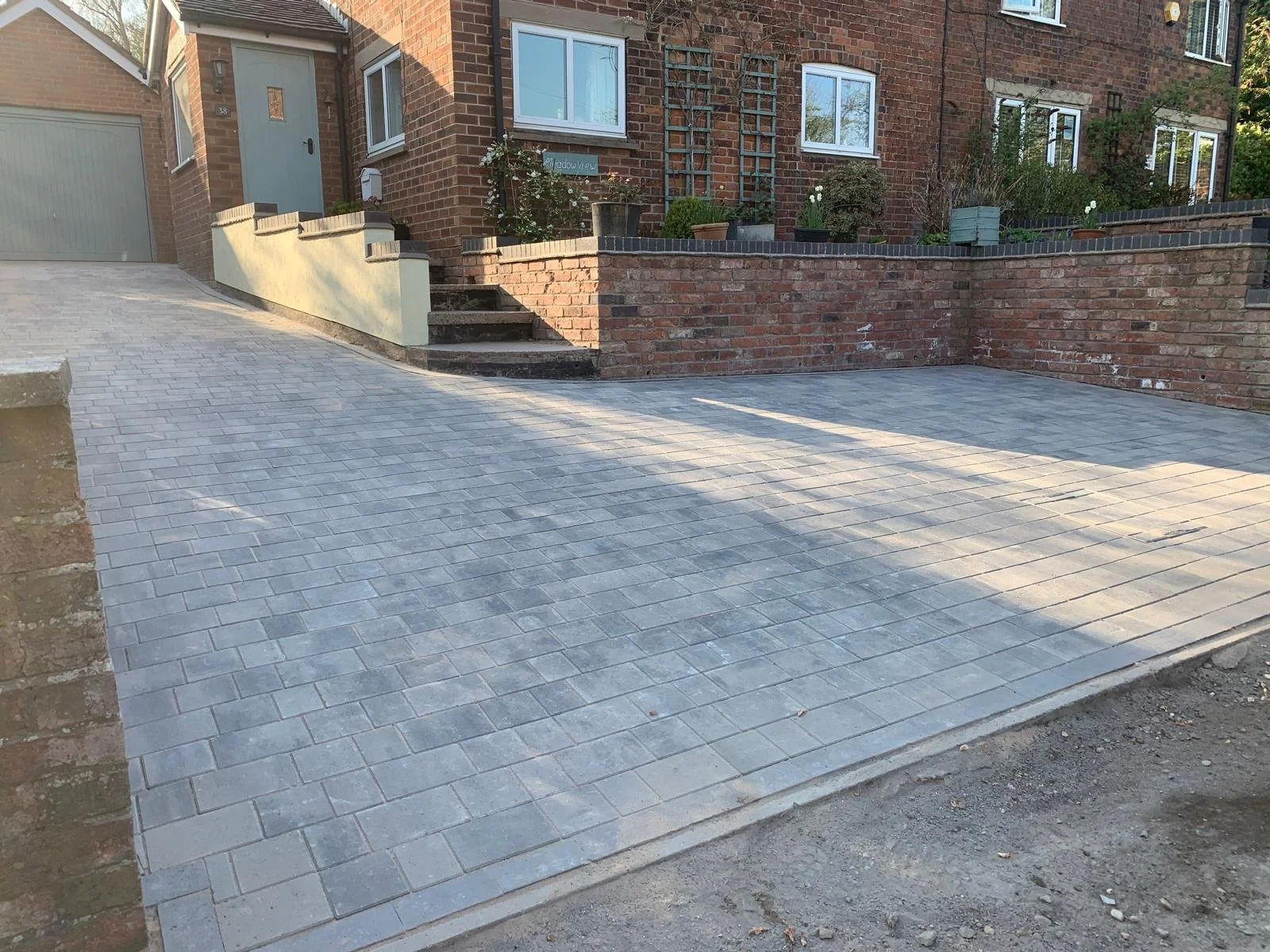 A newly paved driveway with gray interlocking bricks in front of a brick house with stairs and potted plants.