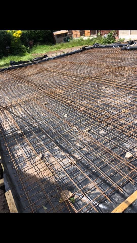 Reinforced concrete foundation prepared with steel rebar mesh on black plastic drainage sheet, outdoors near greenery and buildings.