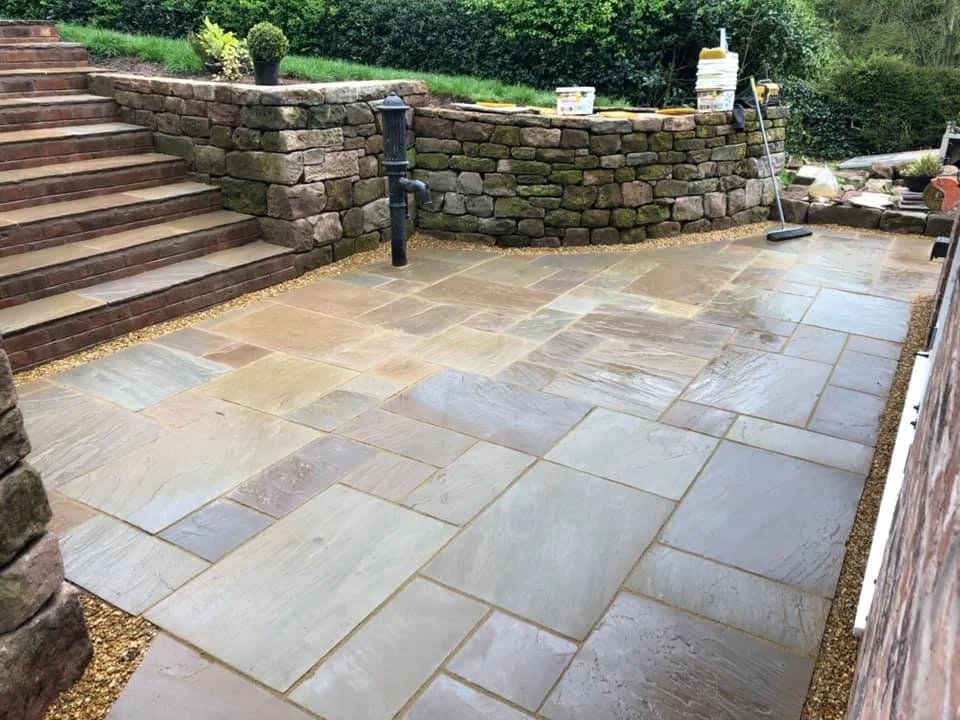 A newly installed stone patio with large rectangular and square stones, bordered by a brick retaining wall and steps on the left, with gardening tools and supplies in the background.