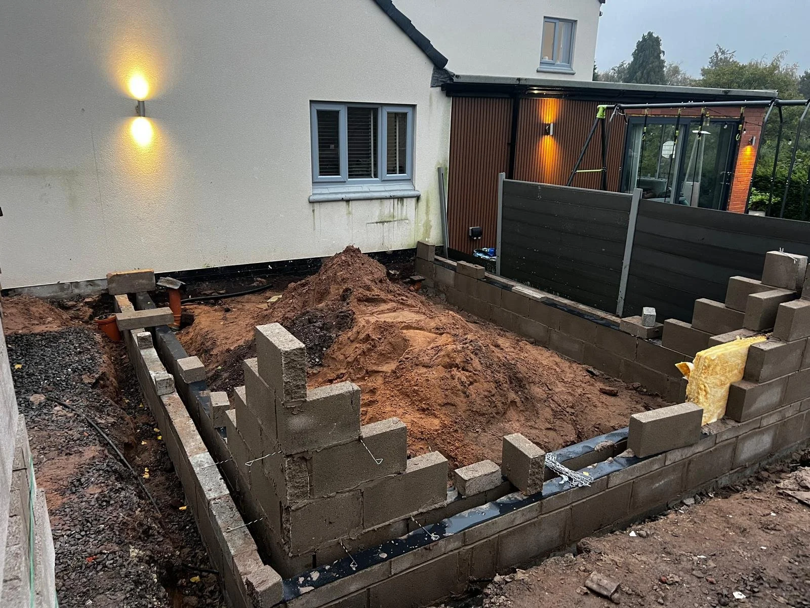 Construction of a small brick wall and foundation in a backyard with dirt piles and building materials.