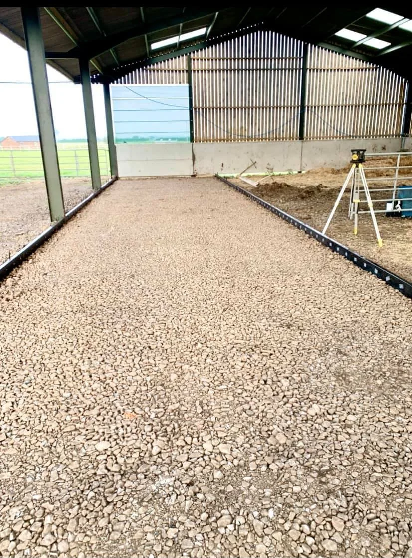 Newly constructed gravel flooring inside a barn or shed with a metal roof, wooden slat walls, and a level concrete wall at the far end, with construction tools nearby.