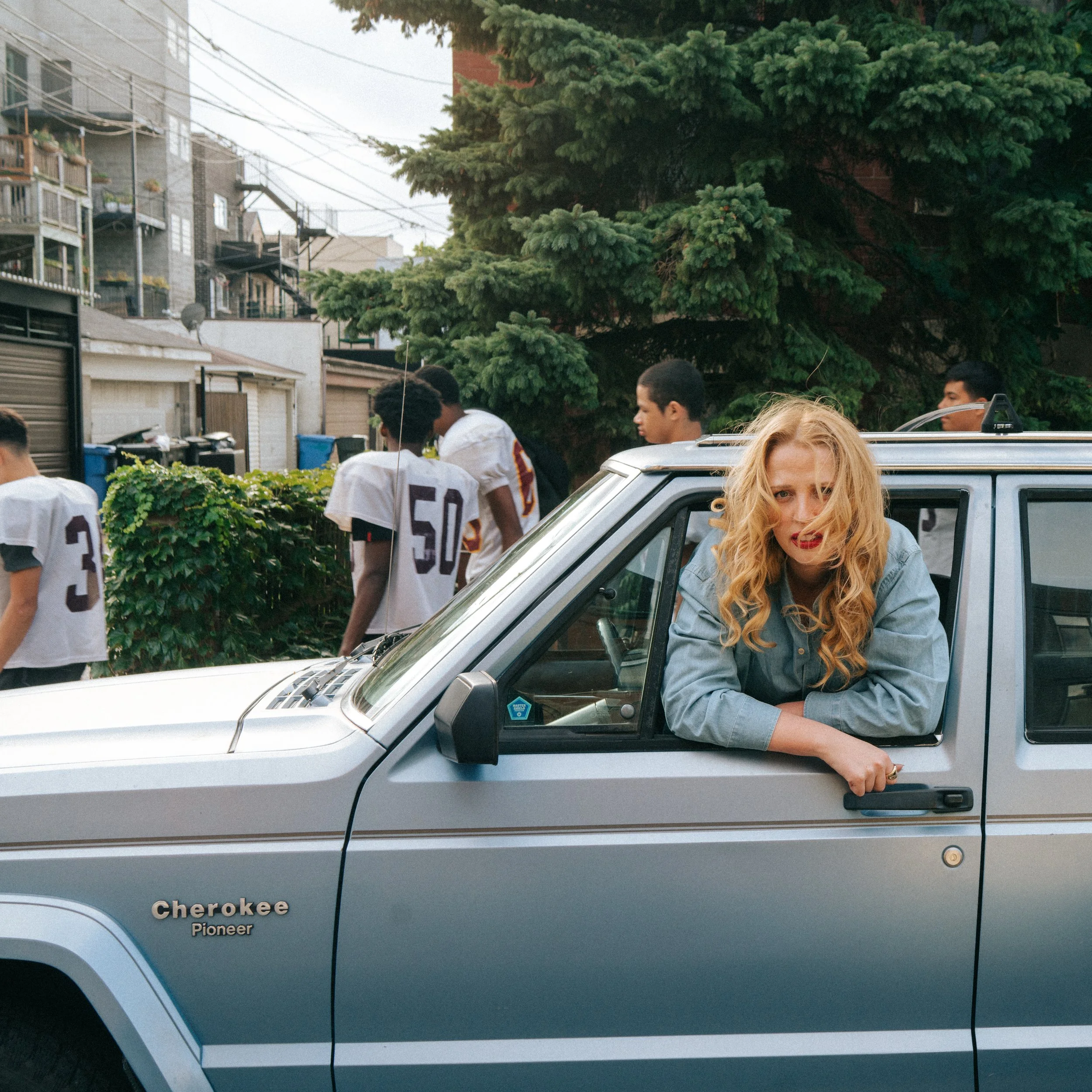 Elizabeth Moen wearing denim in an old, boxy car - a team of high school football players walks by.