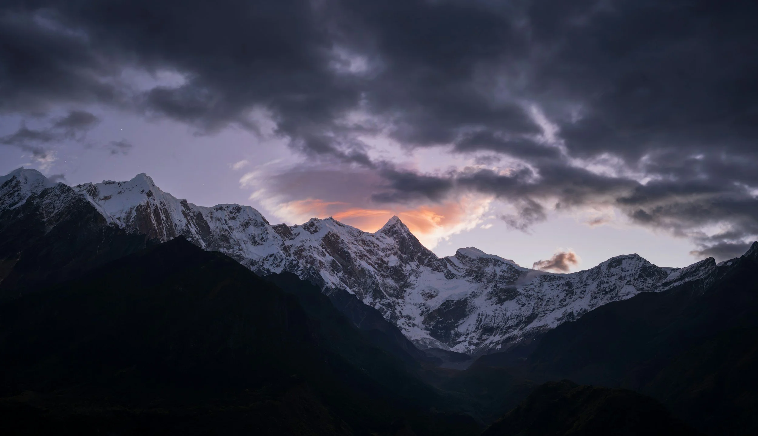 Snow-capped mountains under a dark, cloudy sky at sunset or sunrise.
