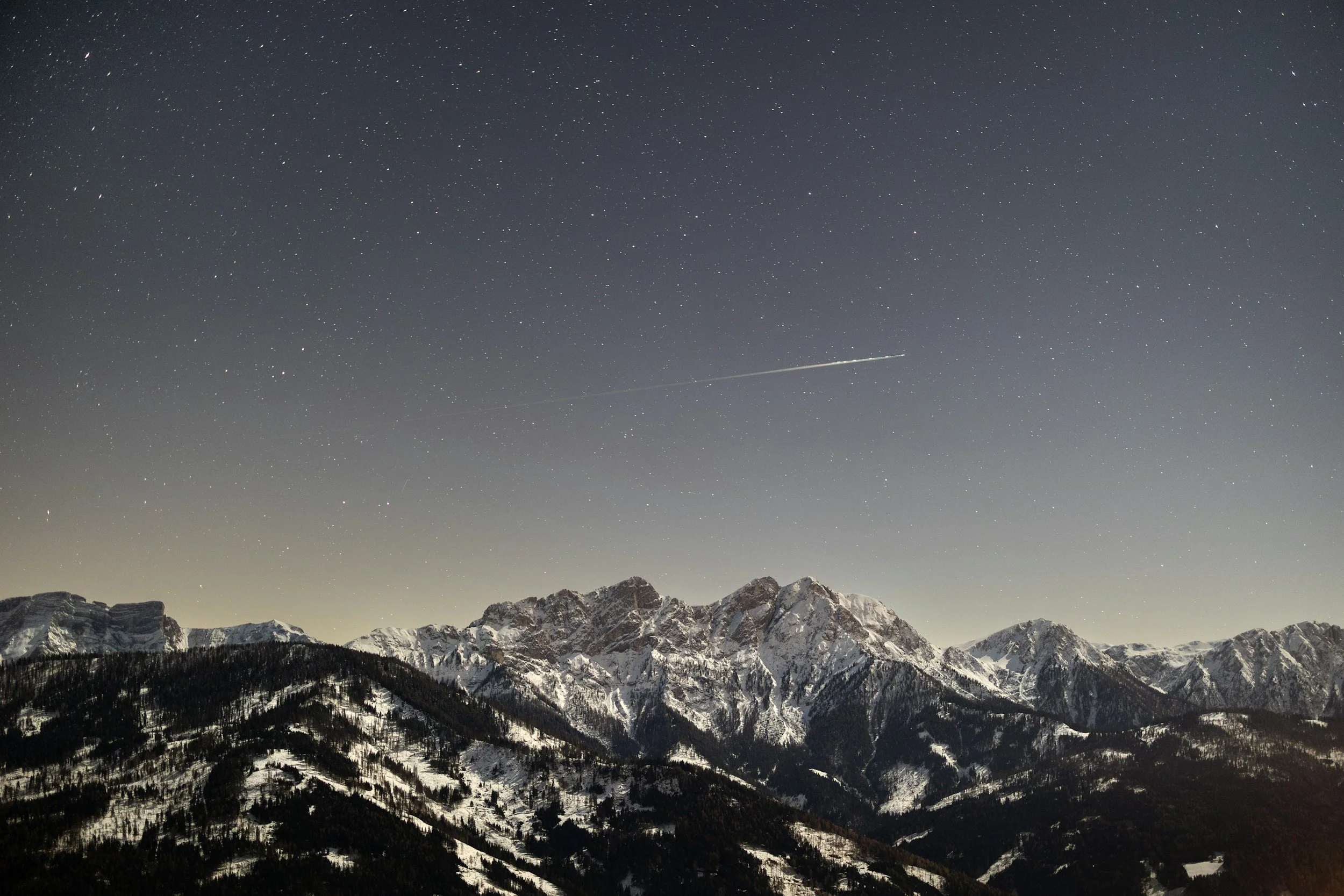 Nighttime mountain landscape with star-filled sky and a visible shooting star or airplane trail.