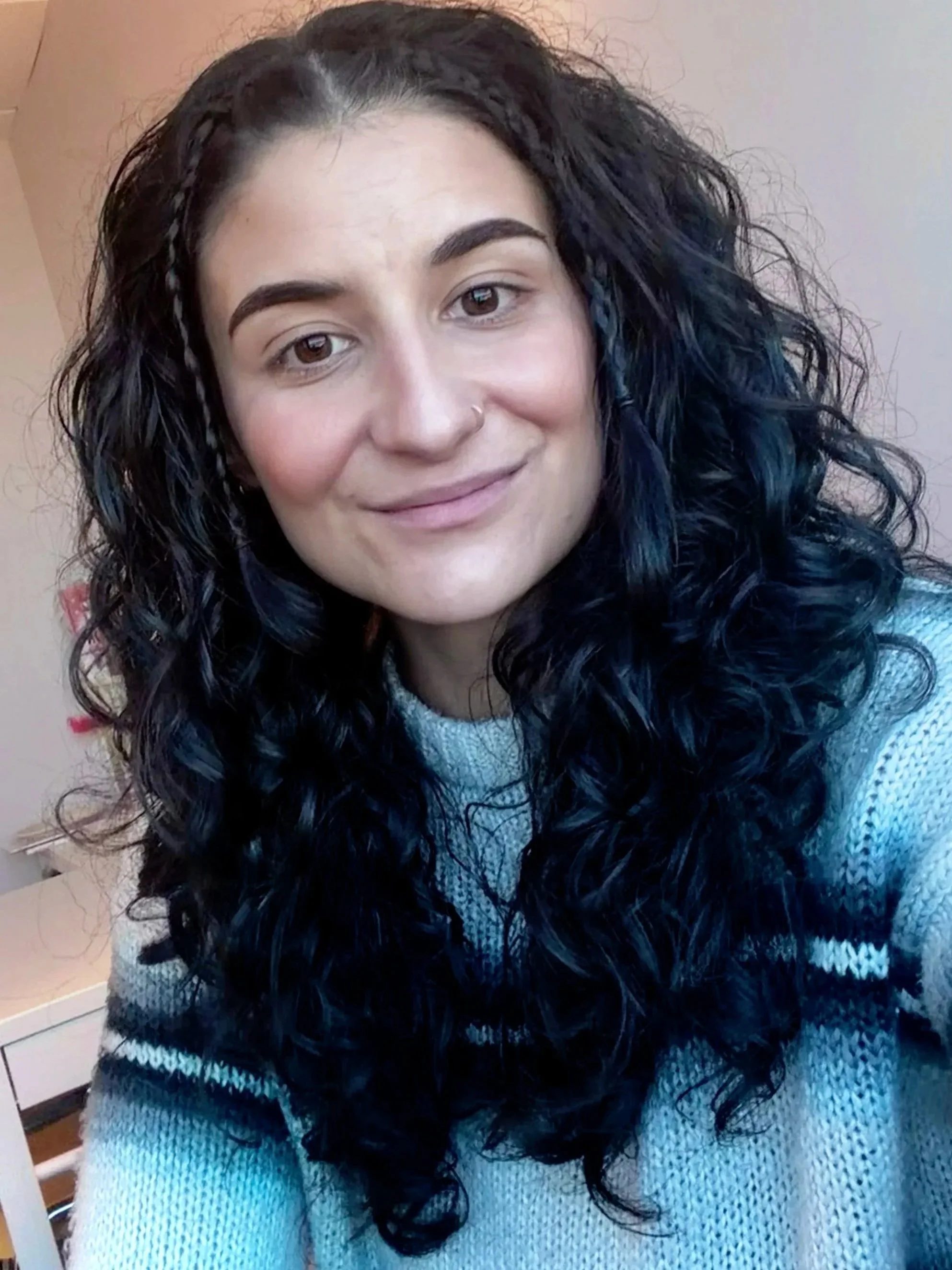 A woman with long, curly black hair, light skin, and dark eyebrows wearing a gray knitted sweater.