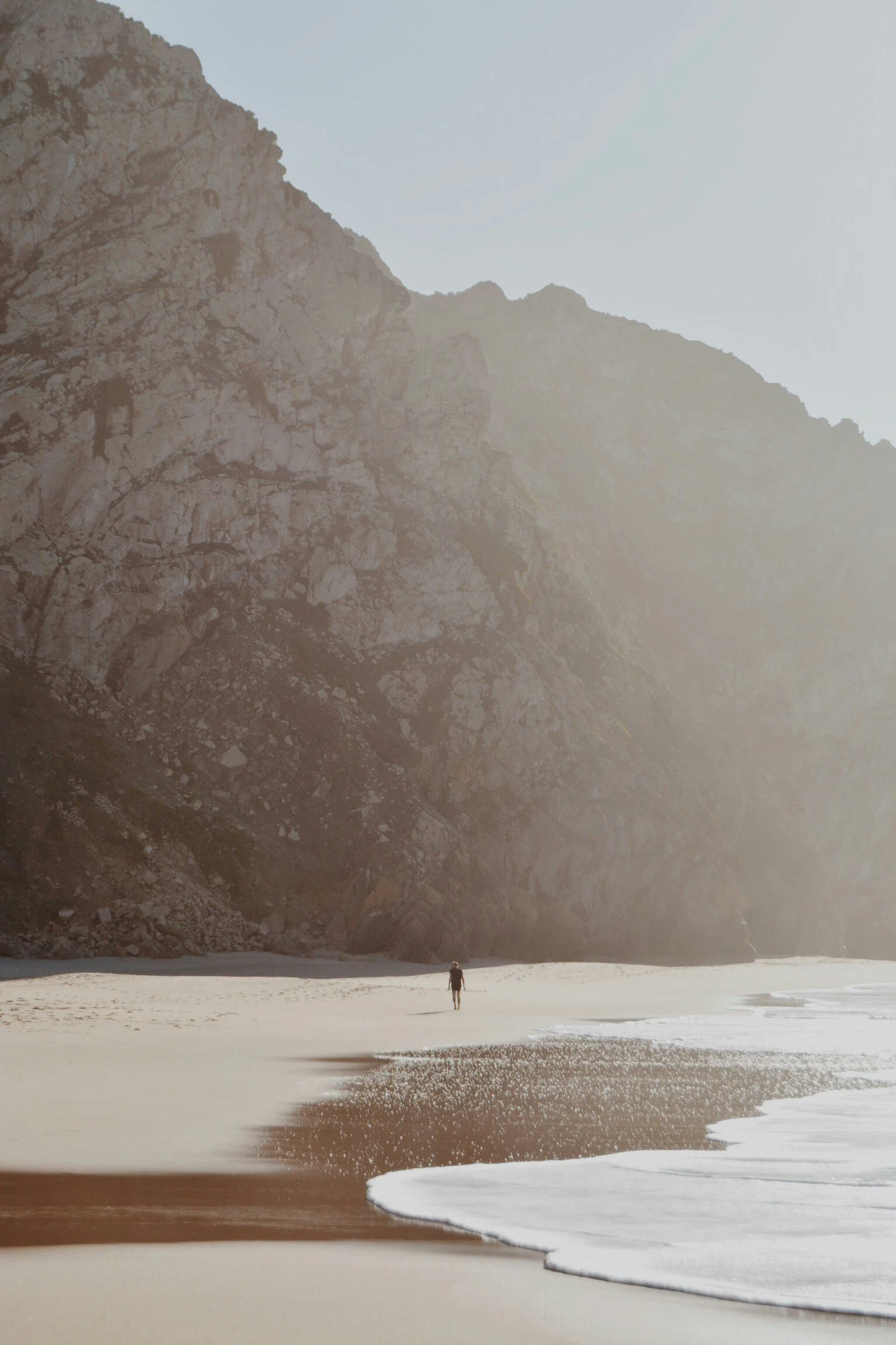 A person standing on a sandy beach with ocean waves gently approaching, with rocky cliffs in the background and a mostly clear sky.