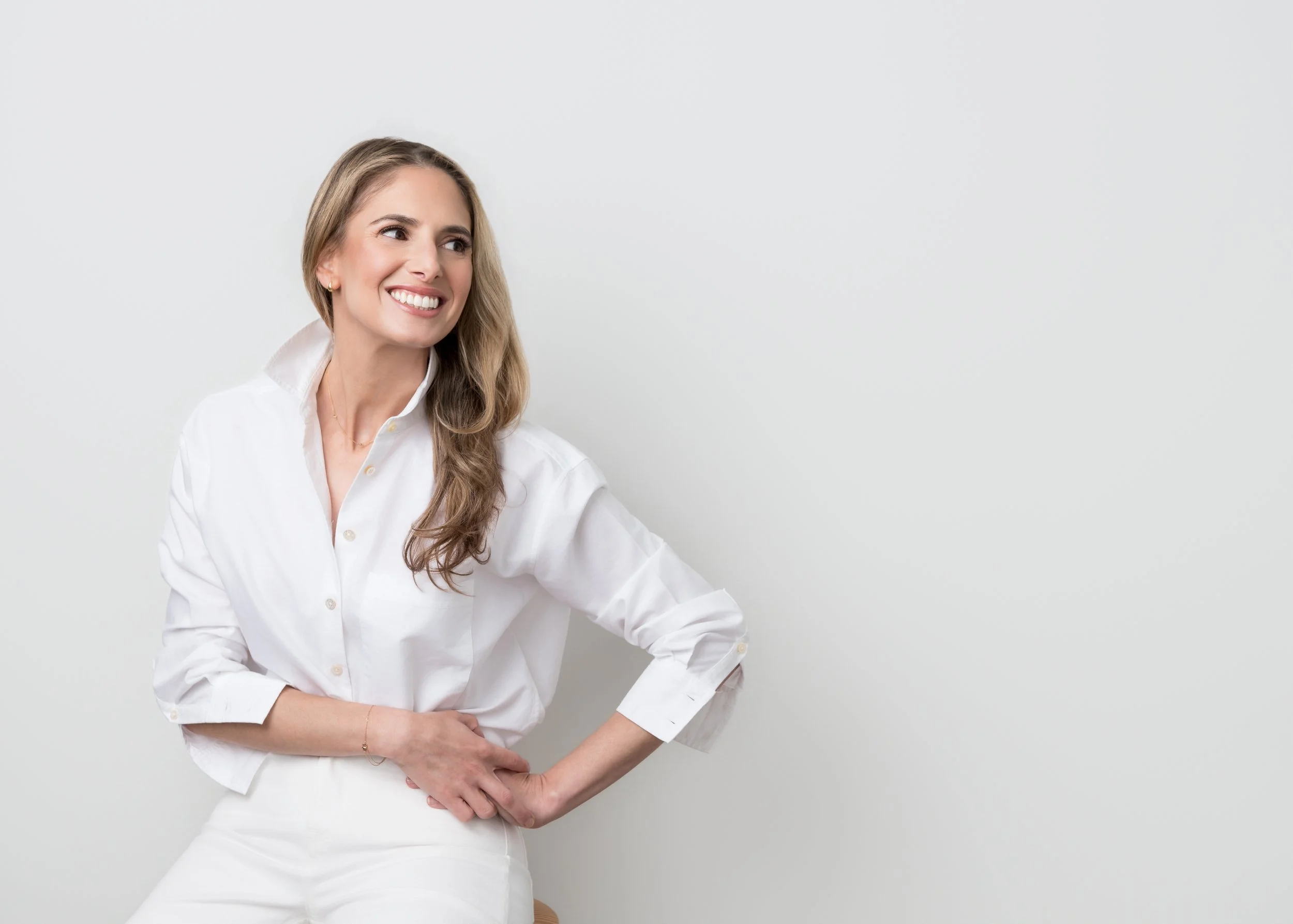 A smiling woman with long blonde hair wearing a white shirt, standing against a plain light gray background.