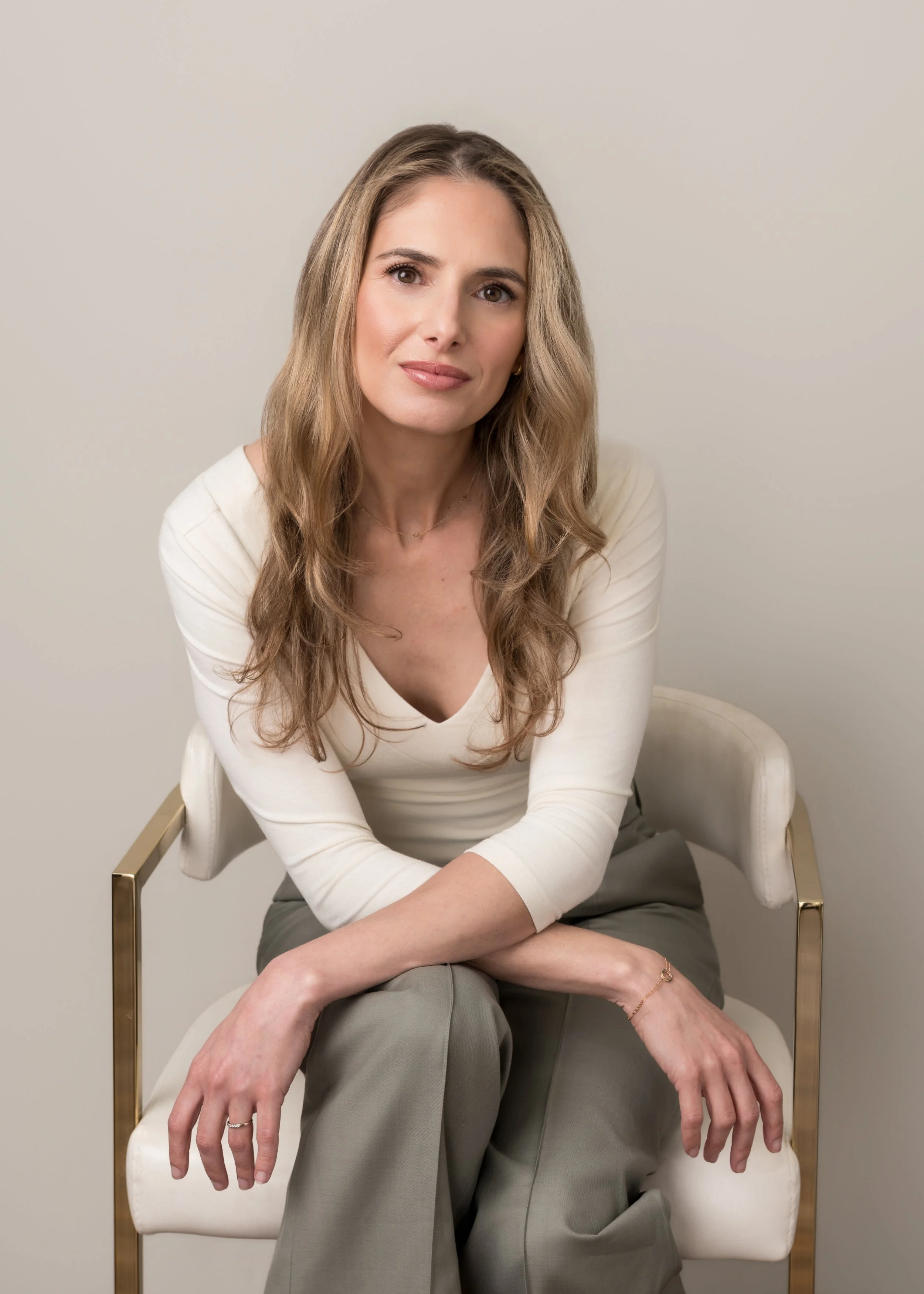 A woman sitting on a white chair with gold armrests against a plain beige background. She has long, wavy light brown hair, and is wearing a cream-colored top and gray pants. She is looking at the camera with a slight smile, her hands resting on her lap, and wearing minimal jewelry.