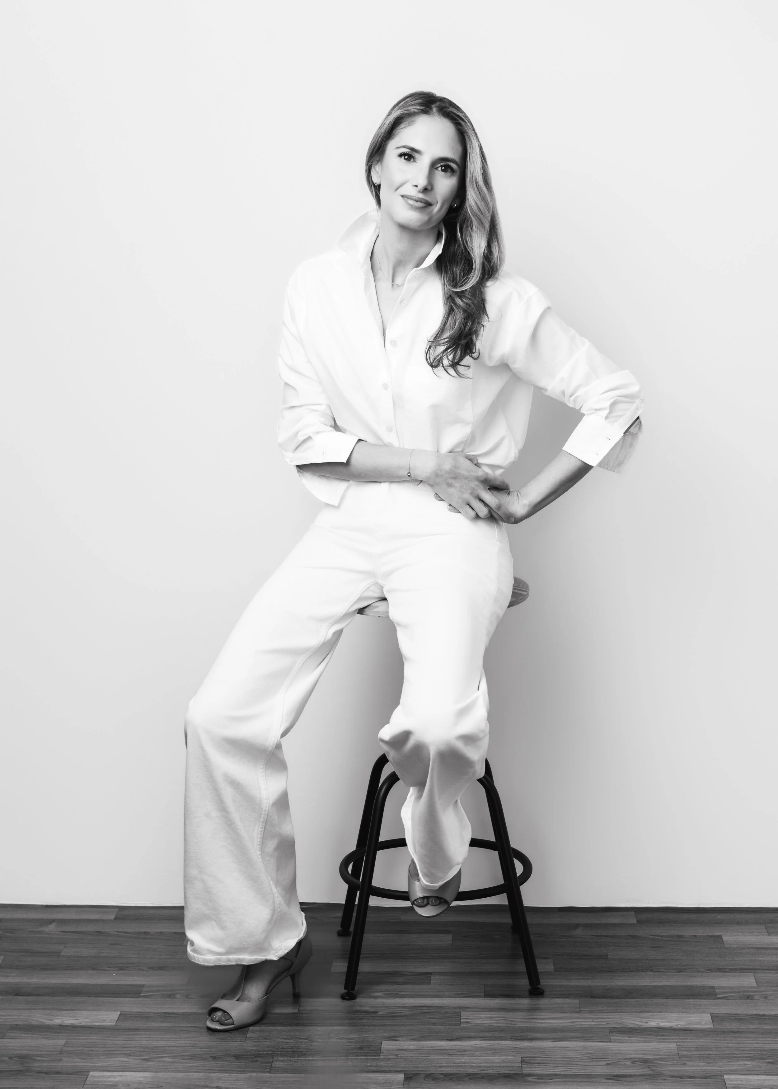 Black and white photo of a woman sitting on a stool against a plain wall, with one arm on her hip and the other resting on the stool, wearing a white shirt, white pants, and high-heeled shoes.