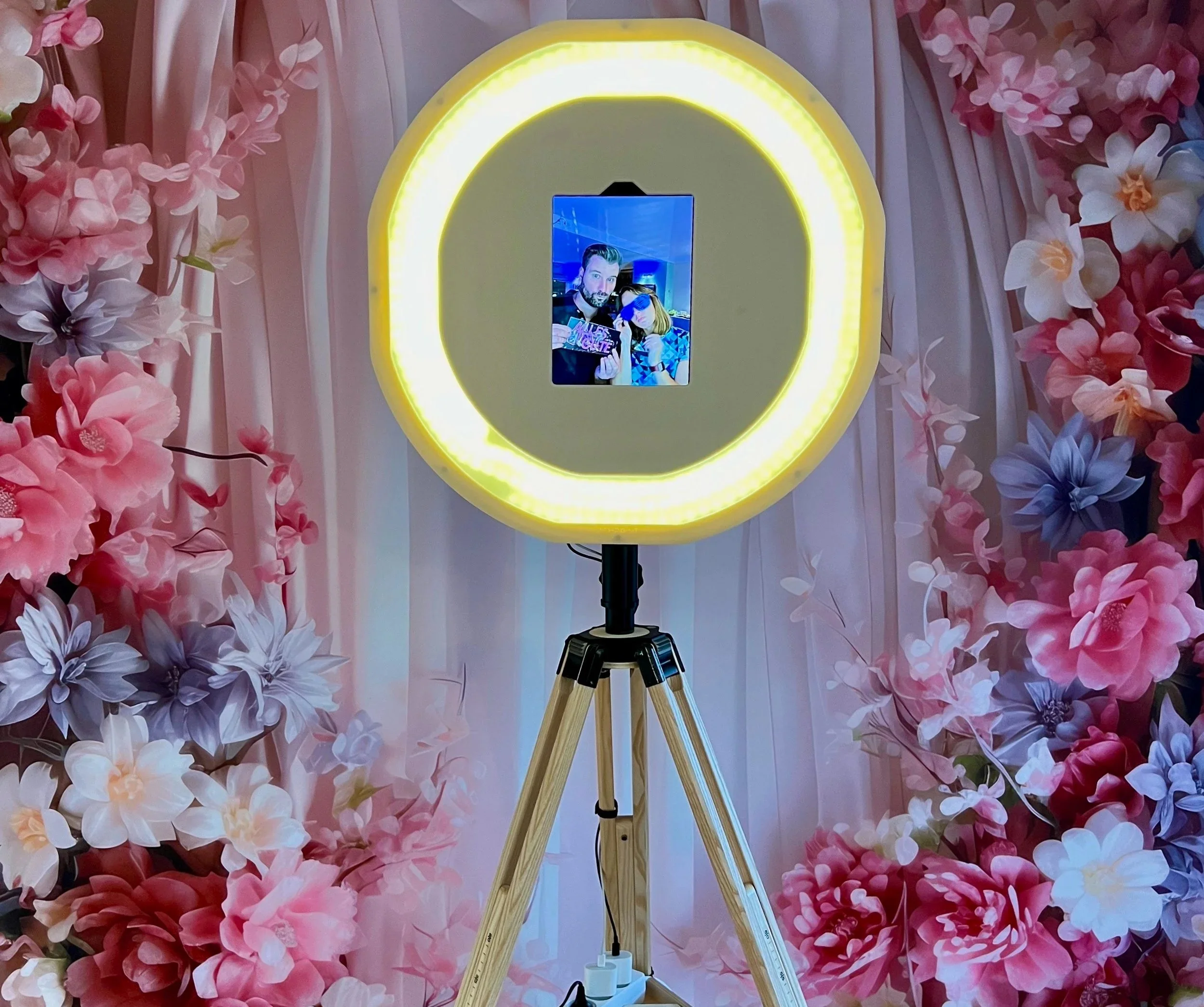Die Social Fotobox beleuchtet mit großem iPad Display. Dahinter ein Hintergrund mit Blumen.